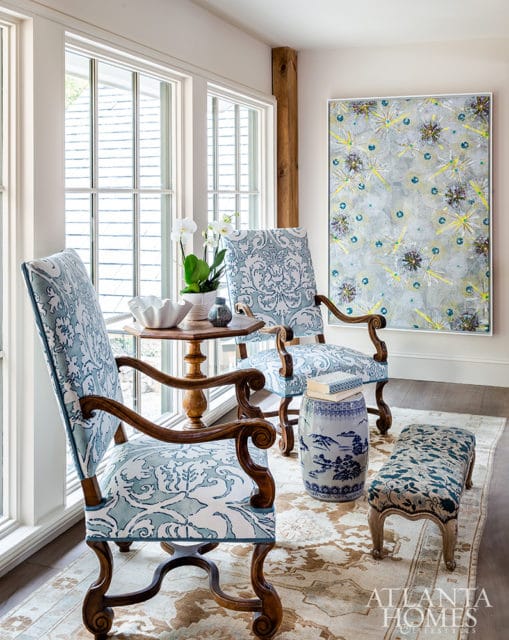 Two upholstered armchairs with blue and white damask pattern, wooden side table with orchids, and a decorative garden stool in a room with large windows and abstract art.