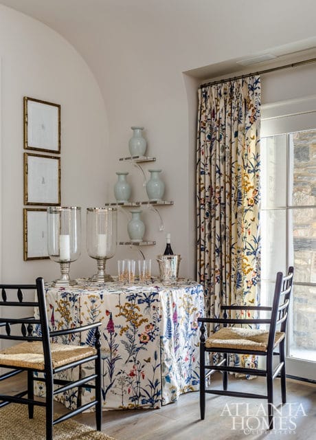 Dining area with a round table covered in a floral print tablecloth, two chairs with woven seats, and curtains with a matching floral pattern. Vases are displayed on shelves on the wall.