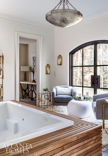 White soaking tub with wooden slatted surround, gray armchairs, arched window with forest view, and ornate chandelier.