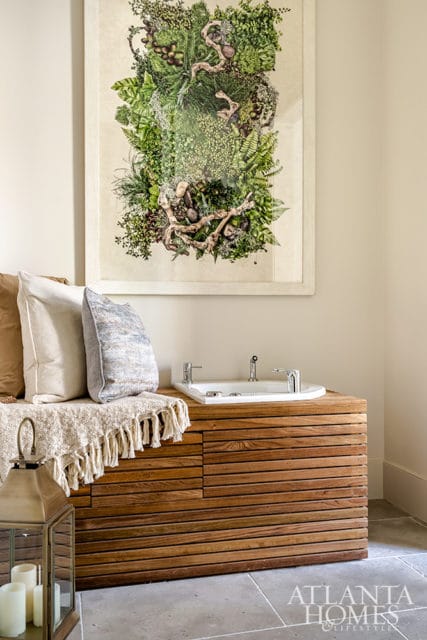 Bathroom with a white soaking tub set into a slatted wooden vanity, a textured throw blanket with tassels, and a large framed botanical artwork on the wall.