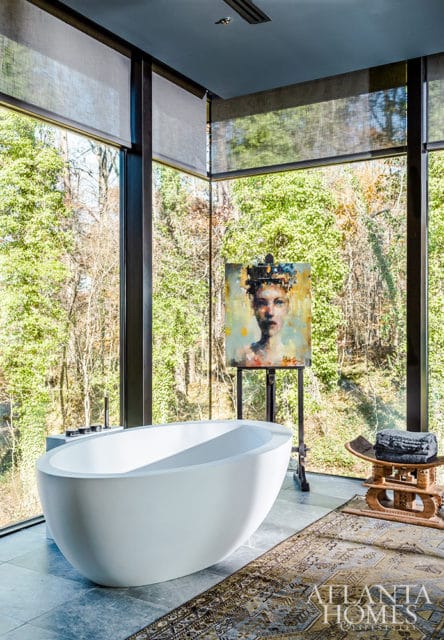 Modern bathroom with a white freestanding bathtub, large windows overlooking trees, and a painting of a woman on an easel.
