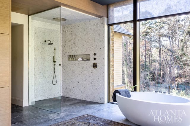 Modern bathroom with a freestanding white bathtub, glass-enclosed shower with terrazzo walls, and large windows overlooking a wooded area.