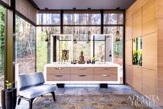 Modern bathroom with a floating wood vanity, dual sinks, and large windows overlooking a forest. A Buddha statue and artwork adorn the vanity, with a grey upholstered chair to the left.