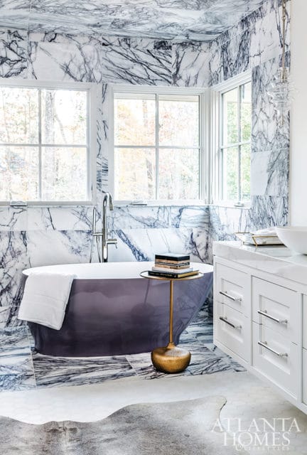Purple freestanding bathtub in a bathroom with dramatic marble walls and ceiling, white vanity, and a gold side table stacked with books.