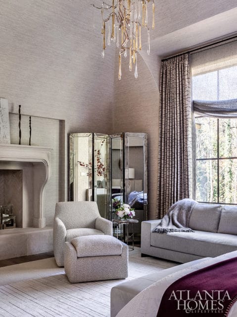 Bedroom with textured grey wallpaper, a stone fireplace, mirrored screen, armchair with ottoman, grey sofa with throw blanket, and a large window with patterned curtains.