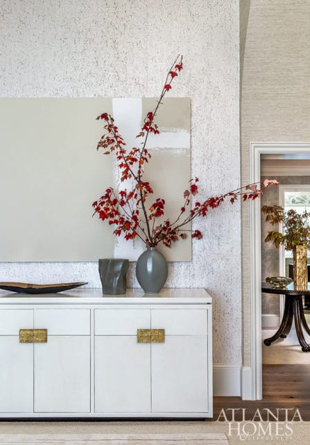White credenza with ornate brass handles topped with a grey vase holding red-leafed branches and abstract art on textured wallpaper.