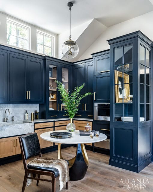 Navy blue kitchen cabinetry with brass hardware, marble countertops and backsplash, and light wood lower cabinets. A round table with a gold base sits in the center with a vase of greenery.
