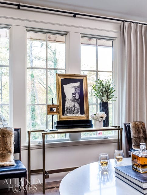 Living room with large windows, a gold-framed abstract artwork on a console table, and two chairs with fur throws.