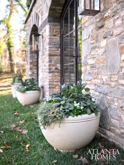 Stone exterior wall with arched windows and large planters filled with greenery and herbs on a grassy lawn.
