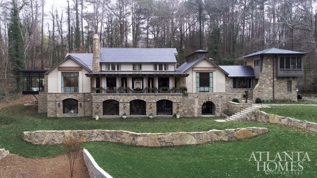 Large stone and clapboard house with multiple wings, a covered balcony, arched windows, and a glass-enclosed sunroom, set against a backdrop of wooded trees.
