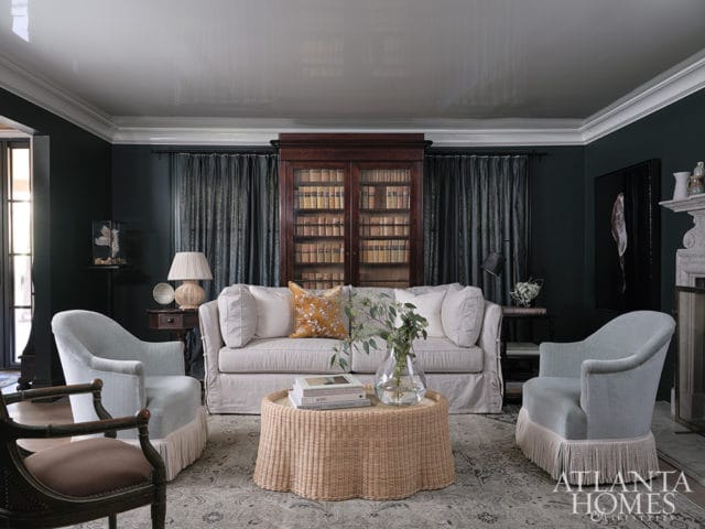 Living room with dark green walls, a slipcovered sofa, two light blue armchairs with fringe, a wicker coffee table, and a large wooden bookcase filled with books.