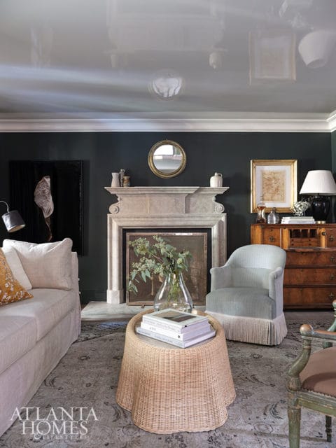 Living room with dark green walls, a stone fireplace, a light-colored sofa, a fringed armchair, and a wicker coffee table topped with books.