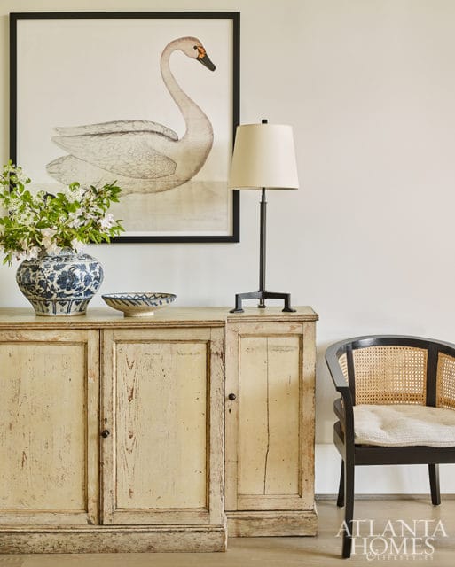 Distressed cream sideboard with a framed swan print above, a blue and white vase with flowers, a lamp, and a black cane-backed chair