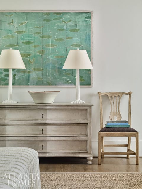 Light gray dresser with three drawers topped with two white lamps and a large framed artwork of a pond with lily pads. A wooden chair with books stacked on the seat sits to the right.
