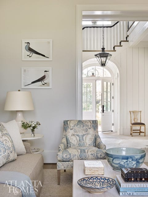 Living room with a neutral sofa, armchair upholstered in blue and white floral fabric, and a coffee table with decorative bowls and books. Two framed bird prints hang on the wall.