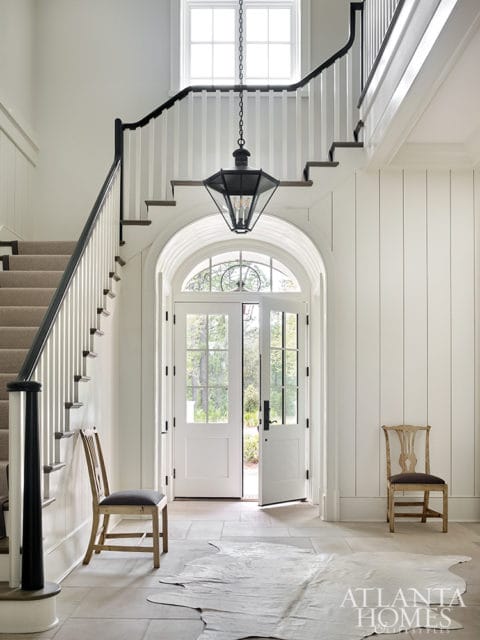 Foyer with a grand staircase, black railing, carpeted stairs, and a large black lantern chandelier. Double doors open to the outside, with two wooden chairs and a cowhide rug on the stone floor.