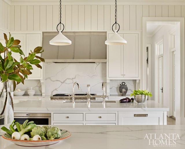 White kitchen with paneled walls, marble countertops and backsplash, stainless steel range hood, and two white pendant lights over the island.