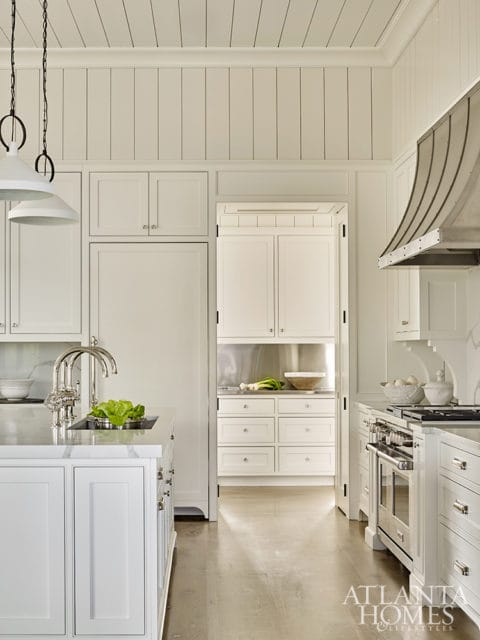 White kitchen with paneled walls and ceiling, marble island with sink, stainless steel range with curved hood, and view into pantry with white cabinetry and stainless steel backsplash.