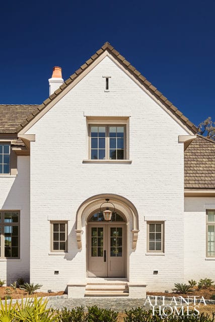 White brick house with arched entryway, double doors, and multiple windows under a brown shingle roof.