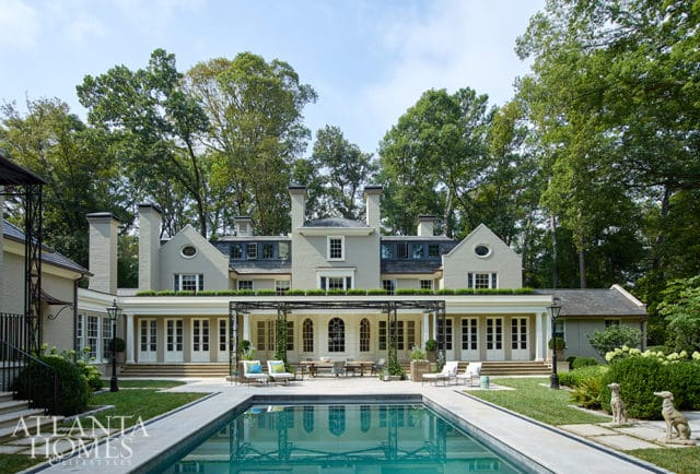 Light grey brick home with multiple chimneys, dormer windows, and a covered patio overlooking a swimming pool and manicured lawn.