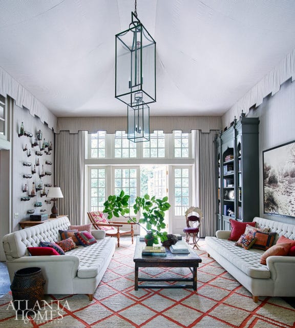 Living room with two tufted sofas, a dark wood coffee table with a large plant, and a large geometric rug. A tall dark green bookshelf is on the right wall.
