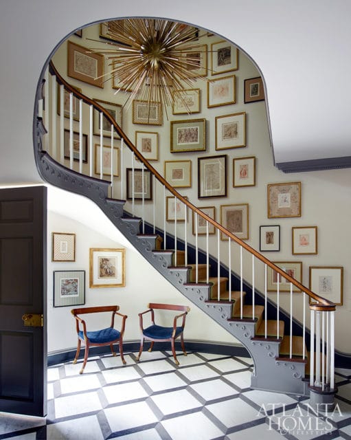 Curved staircase with gray railing and white spindles, adorned with a gallery wall of framed art and a starburst chandelier. Two blue upholstered chairs sit on a black and white geometric tiled floor.