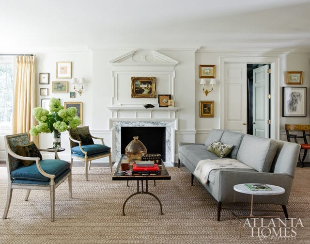 Living room with a gray sofa, two blue armchairs, a fireplace with a marble surround, and a woven rug. Artwork adorns the walls, and a large vase of green flowers sits on a side table.