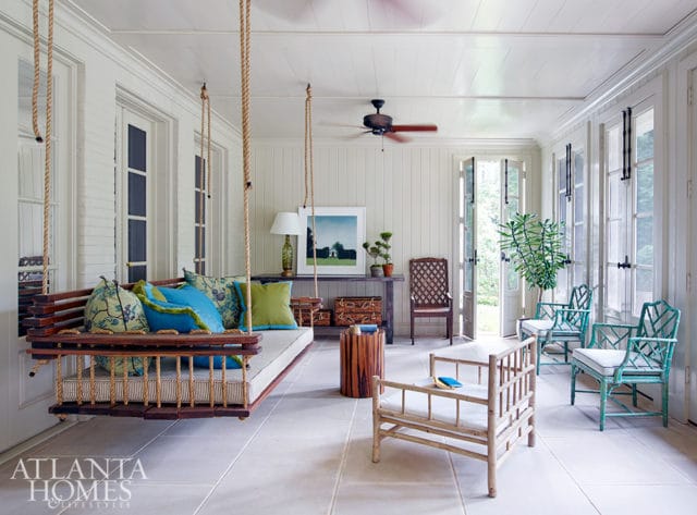 Sunroom with a wooden swing bed, light-colored paneled walls, and large windows. Decorated with colorful pillows, bamboo furniture, and a ceiling fan.