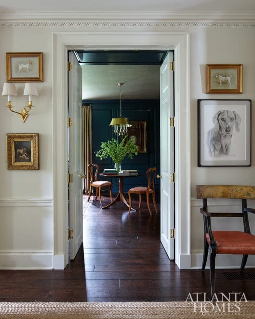 Hallway with white walls and wainscoting, leading to a dark teal room with a round table, two chairs, and a green chandelier. Dog portraits hang on the walls.