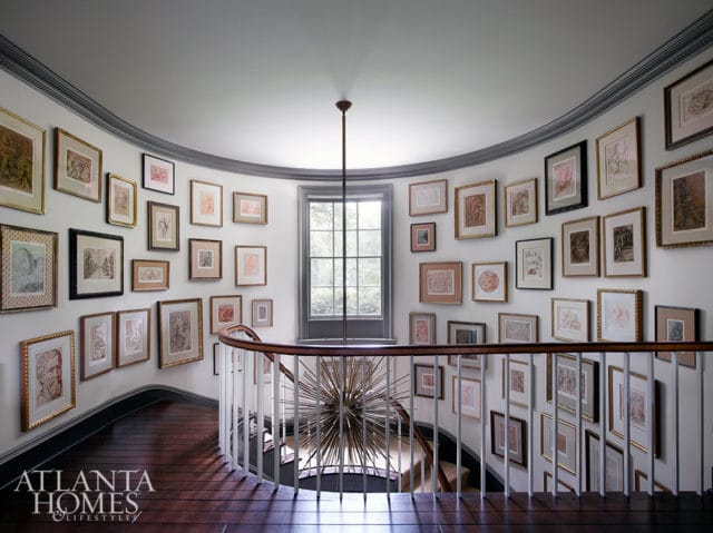 Curved staircase landing with walls covered in framed art, dark wood floors, white railing, and a central starburst chandelier.