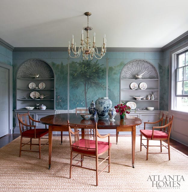 Dining room with oval wooden table, red upholstered chairs, and a mural depicting a tree and landscape on the walls. Built-in shelves display blue and white porcelain.