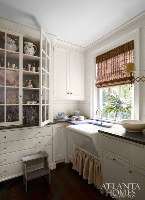 White kitchen with glass-front cabinets displaying silver and ceramic pitchers, a farmhouse sink with a ruffled skirt, and a bamboo window blind.