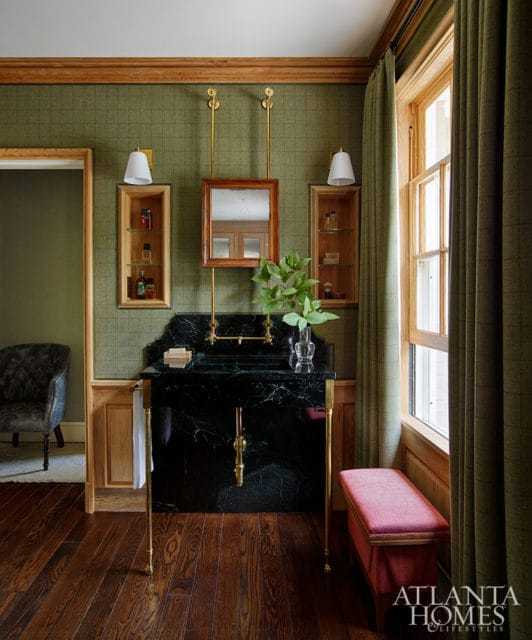 Green tiled bathroom with a black marble vanity, brass fixtures, wooden floors, and a tufted armchair visible through a doorway.