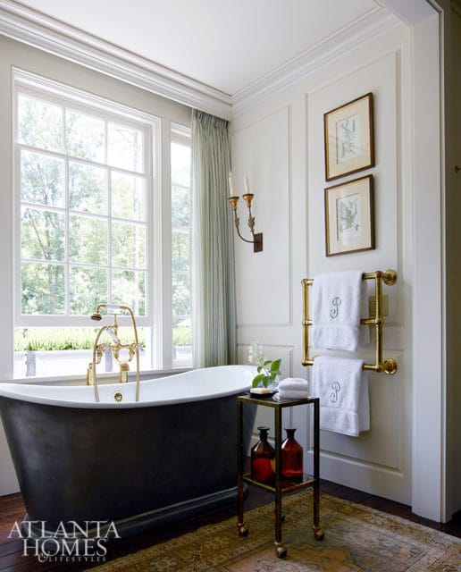 Dark gray freestanding bathtub with brass fixtures next to a window with green striped curtains. A brass towel rack with embroidered towels hangs on the wall.