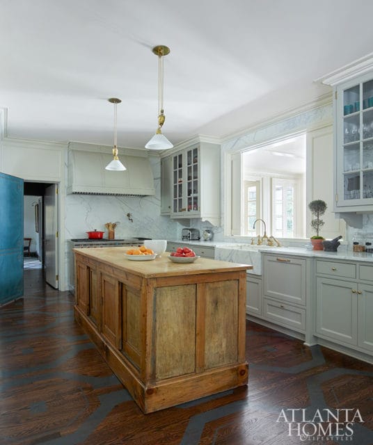 Light gray kitchen with marble backsplash, white farmhouse sink, and a rustic wooden island topped with fruit. Dark wood floors feature a geometric pattern.