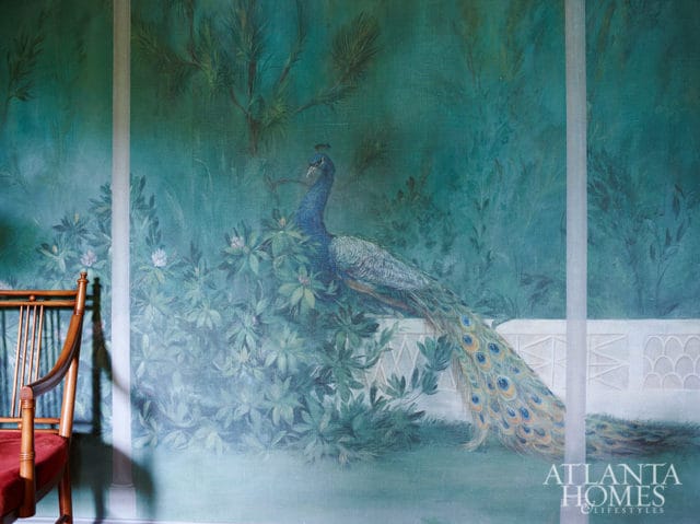 Mural of a peacock amidst lush greenery with a white balustrade, next to a wooden chair with a red cushion.