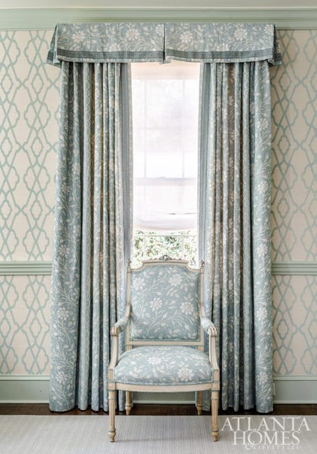 Light blue floral upholstered armchair in front of a window with matching floral curtains and trellis-patterned wallpaper.