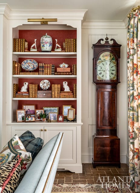 Built-in white shelving unit with red textured wallpaper backing, filled with books, decorative plates, dog figurines, and framed family photos. A grandfather clock stands to the right, next to patterned curtains.