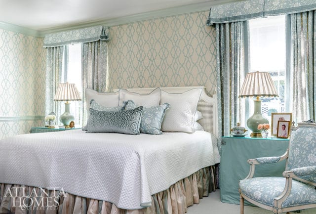 Bedroom with a white quilted bed, patterned wallpaper in cream and light blue, and matching curtains. A light blue armchair with floral upholstery sits to the right.