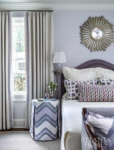 Bedroom with a purple upholstered headboard, white bedding, and patterned pillows. A sunburst mirror hangs on the pale lavender wall above the bed.