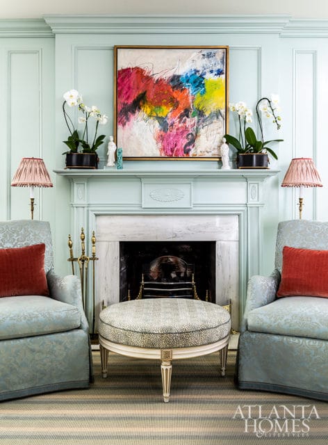 Light blue paneled walls with a marble fireplace, flanked by two upholstered chairs and topped with abstract art and orchids. A patterned ottoman sits in front of the fireplace.