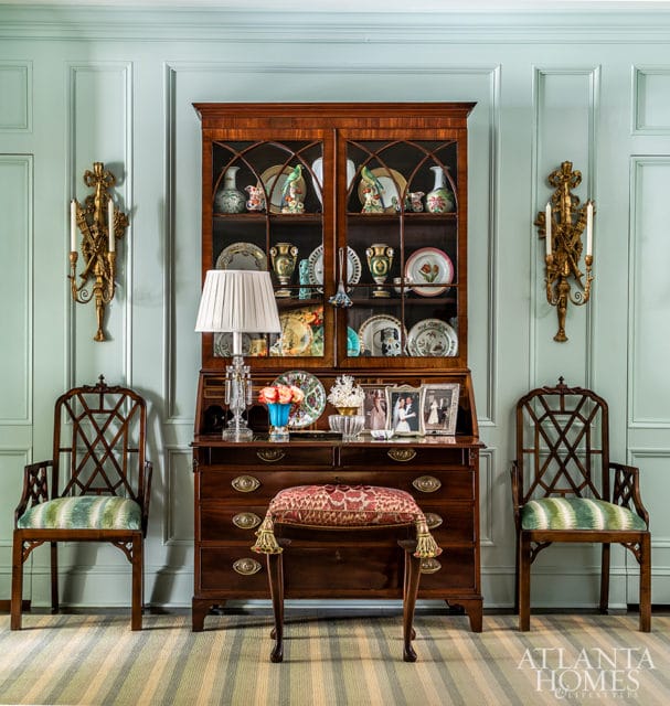 Light blue paneled walls with a wooden secretary desk filled with decorative plates and vases, flanked by two chairs and ornate sconces.