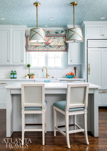 Light blue kitchen with white subway tile backsplash, brass hardware, and a patterned Roman shade above the sink. Two white bar stools with blue seats face a kitchen island.