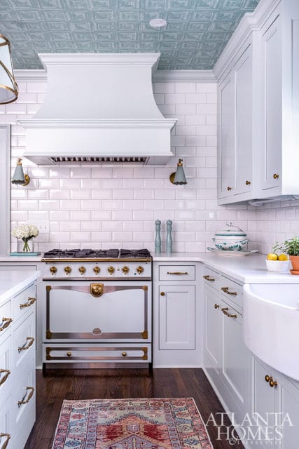 White kitchen with white subway tile backsplash, a white range hood, a vintage-style range with brass accents, and a decorative tin tile ceiling in a light blue hue.