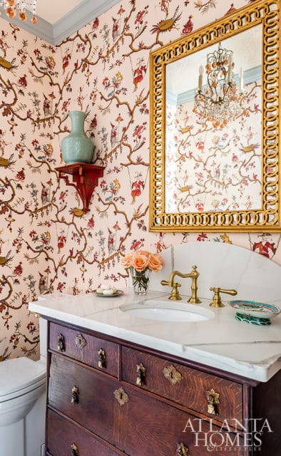 Powder room with Chinoiserie wallpaper, marble countertop, antique wood vanity with brass hardware, and a gold ornate mirror reflecting a crystal chandelier.