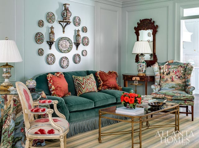 Teal velvet sofa with coral and patterned pillows in a living room with a wall of decorative plates, a mirrored wall sconce, and a wingback chair upholstered in a scenic print.