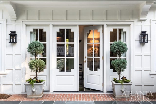 White double doors with geometric glass panes open to reveal a glimpse of a lamp and chair inside. Flanked by topiaries in lion-head planters and black lanterns.