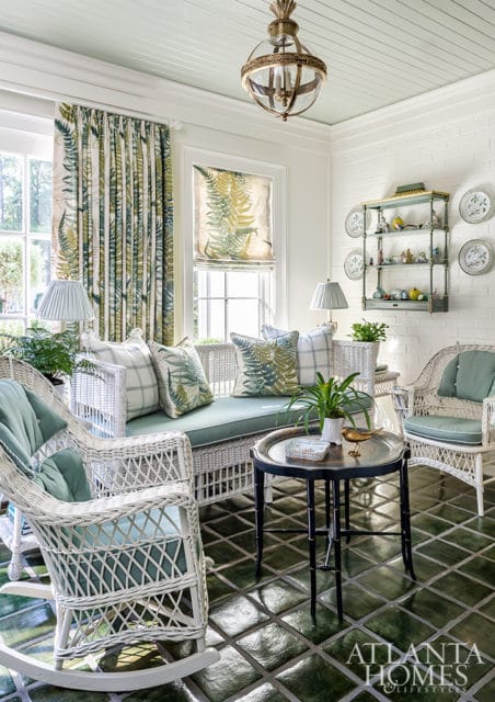 Sunroom with white wicker furniture, green fern-patterned curtains and pillows, and glossy green tile flooring. A brass chandelier hangs from the light blue ceiling.