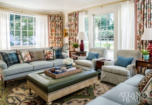 Light blue sofa with patterned pillows, green leather ottoman, and striped armchairs in a living room with large windows and floral curtains.