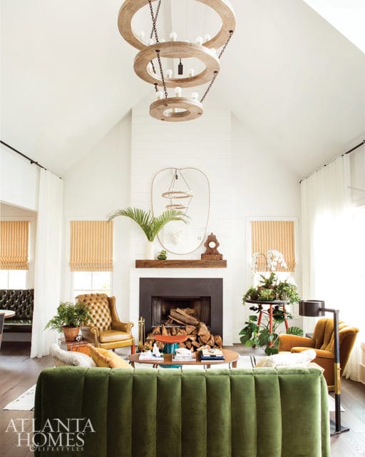 Living room with vaulted ceiling, white shiplap walls, and a dark fireplace. Features a green velvet sofa, tufted leather armchairs, and three tiered wooden chandeliers.